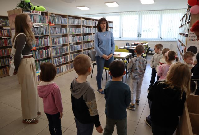 Na zdjęciu znajduje się grupa przedszkolaków, przed nimi stoi jedna kobieta oraz druga po lewej stronie. W tle znajdują się półki z książkami, stoliki z krzesłami oraz okna.