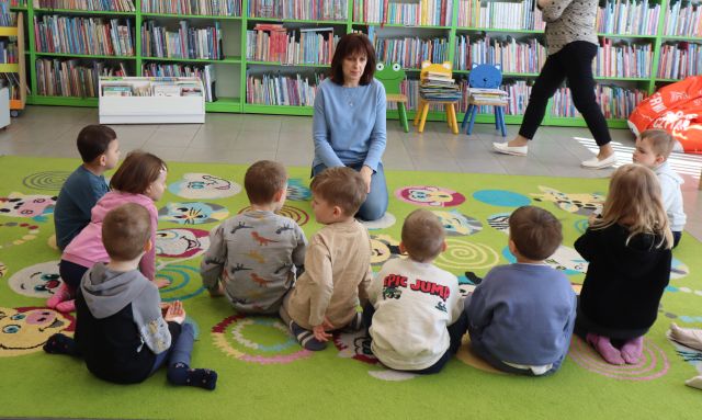 Na zdjęciu znajduje się dziesiątka dzieci siedząca na dywanie przed kobietą, w tle przechodzi inna kobieta oraz znajdują się półki z książkami.
