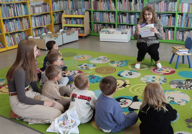 Na zdjęciu znajdują się przedszkolaki oraz kobieta, którzy siedzą na dywanie, a przed nimi siedzi kobieta i pokazuje im naklejki oraz kartę na naklejki.