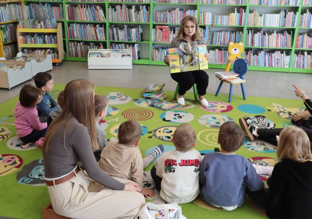 Na zdjęciu znajdują się dzieci siedzące wraz z kobietą na dywanie w półokręgu, a przed nimi siedzi kobieta, która pokazuje im ilustracje w książce. W tle znajdują się półki z książkami.