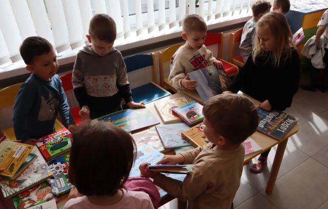 Na zdjęciu znajdują się przedszkolaki stojące przy złączonych stolikach i oglądają wyłożone na nim książki. W tle znajdują się okna.