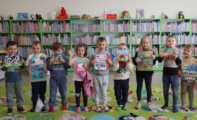 Na zdjęciu znajduje się dziewięcioro przedszkolaków stojących do zdjęcia z książkami. W tle znajdują się półki z książkami.
