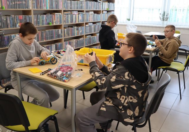 Na zdjęciu znajduje się czwórka chłopców przy dwóch stolikach i układają klocki LEGO. W tle znajdują się półki z książkami.