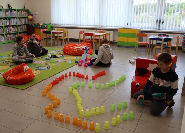 Na zdjęciu znajduje się czterech chłopców, dwójka siedzi na dywanie po lewej, jeden chłopiec układa przy dywanie wieże z kolorowych kubeczków, a czwarty chłopiec układa trasę dla robota z kolorowych kubeczków, za nim znajduje się pociąg z puf. W tle znajduja się okna oraz półki z książkami.