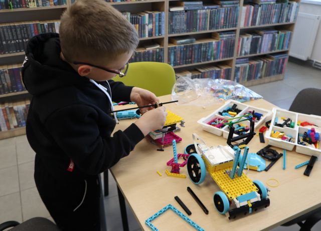 Na zdjęciu znajduje sie układający LEGO przy stoliku chłopiec. Na stoliku stoi plastikowe opakowanie z klockami LEGO oraz samochód. W tle znajdują się półki z książkami.