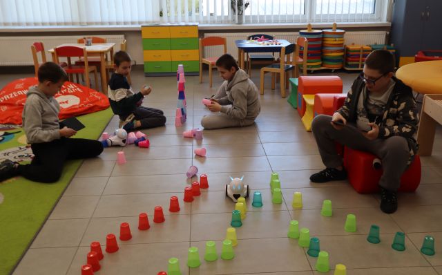 Na zdjęciu znajduje się czwórka chłopców, dwójka układa przy dywanie wieże z kolorowych kubeczków, a jeden jeździ robotem przez zbudowany wcześniej labirynt z kubków. W tle znajdują się stoliczki, szafka oraz okna.