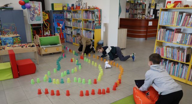 Na zdjęciu znajduje się trójka chłopców, którzy pokonują tor z kubeczków robotami. W tle znajdują się półki z książkami.