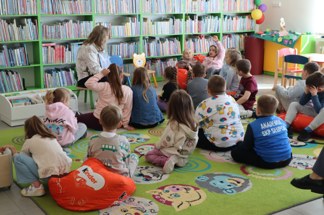 Na zdjęciu znajduje się grupa przedszkolaków siedząca na dywanie i na czerwonych pufach, przed nimi siedzi kobieta i czyta im książkę. W tle znajdują się półki z książkami.