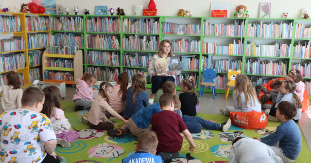 Na zdjęciu znajdują się przedszkolaki siedzące na dywanie, a przed nimi siedzi kobieta i pokazuje im ilustracje książki. W tle znajdują się półki z książkami.