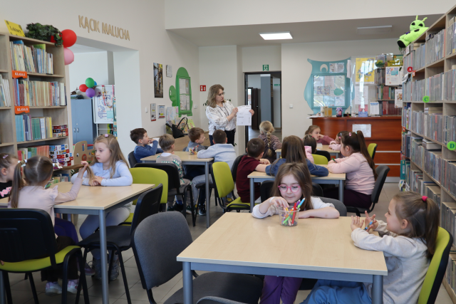 Na zdjęciu znajdują się przedszkolaki przy stolikach, na których leżą pudełka z kredkami. Przed stołami stoi kobieta i pokazuje im kartę pracy. Po bokach znajdują się półki z książkami, a w tle na ścianach rysunki i drzwi.