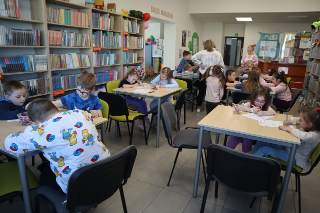 Na zdjęciu znajdują się przedszkolaki przy sześciu stołach i wykonują karty pracy, w tle między stolikami stoja trzy kobiety. W tle znajdują się półki z książkami.