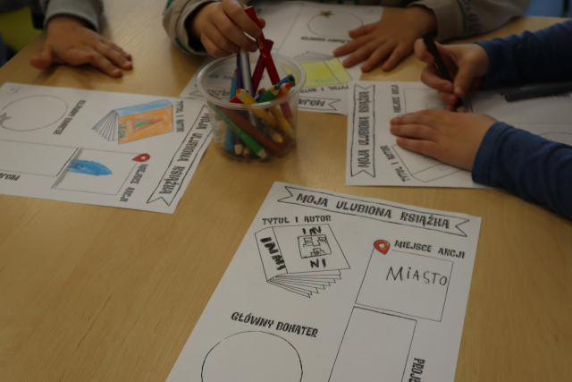 Na zdjęciu znajduje się stolik, na którym znajdują się kredki oraz cztery karty pracy, które są kolorowane przez trójkę dzieci siedzącą przy stole.