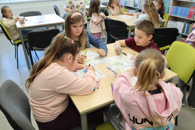 Na zdjęciu znajduje się trójka dzieci przy stoliku i kolorują karty pracy. W tle znajdują się inni uczestnicy zajęć przy stolikach.