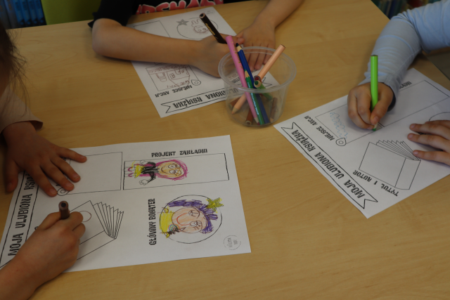 Na zdjęciu znajduje się stół, na którym leżą kredki oraz trzy karty pracy, które są kolorowane przez trójkę przedszkolaków.