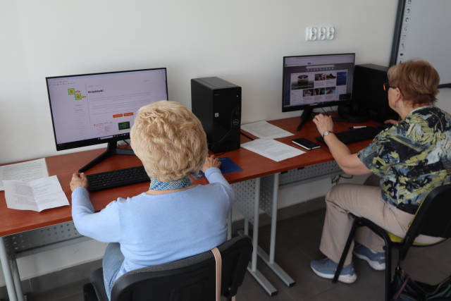 Na zdjęciu znajdują się dwie kobiety siedzące przy biurkach z komputerami i wyszukują informacji w internecie. Warsztaty odbywają się w sali komputerowej.