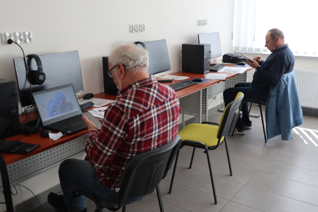 Na zdjęciu znajduje się dwójka mężczyzn przy komputerach, między nimi jest jedno miejsce wolne. Warsztaty odbywają się w sali komputerowej.