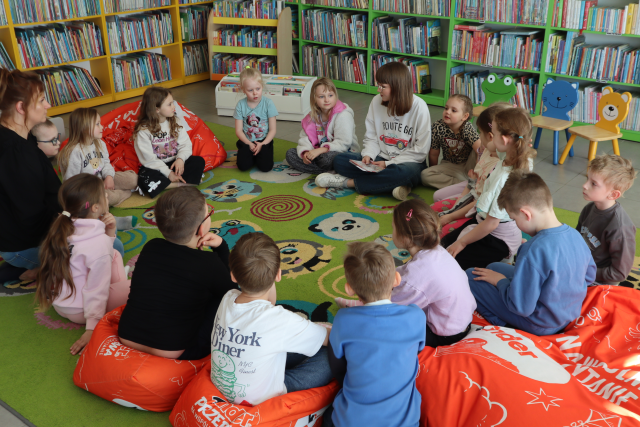 Na zdjęciu znajduje się grupa przedszkolaków wraz z dwoma kobietami na dywanie. W tle znajdują się półki z książkami.