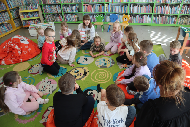 Na zdjęciu znajdują się przedszkolaki siedzące na dywanie, między nimi siedzi kobieta z książką na kolanach. W tle znajdują się półki z książkami.