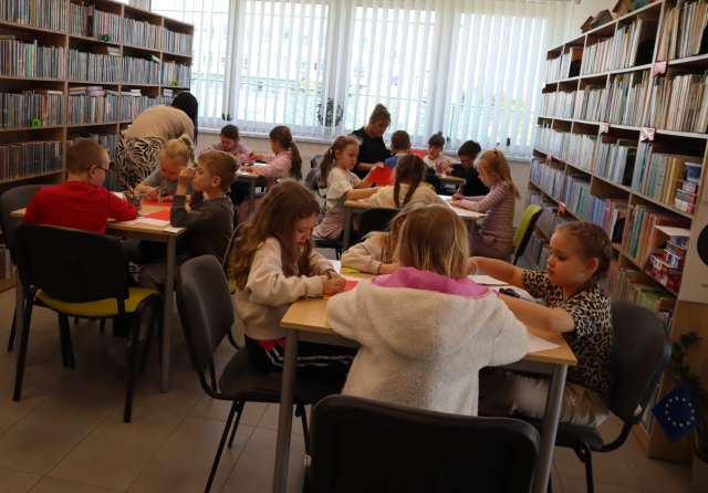 Na zdjęciu znajdują się przedszkolaki przy pięciu stołach. W tle znajdują się okna, a po lewej i prawej stronie półki z książkami.