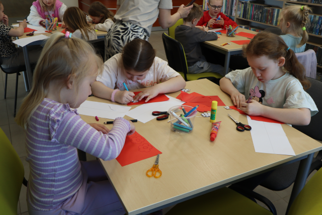 Na zdjęciu znajdują się trzy dziewczynki siedzące przy stoliku i odrysowujące serduszka na czerwonej kartce. Na stoli znajdują się również białe kwiatki i kredki. W tle znajdują się inni uczestnicy zajęć przy stolikach.