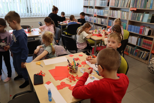 Na zdjęciu znajdują się przedszkolaki przy czterech stolikach, przy których wykonują flagę Polski. Przy jednym stoliku siedzi kobieta. W tle znajdują się półki z książkami oraz okna.