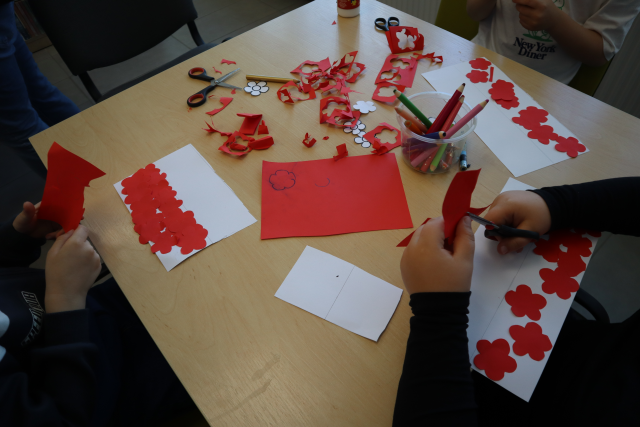 Na zdjęciu znajduje się stolik, przy którym przedszkolaki wykonują biało czerwoną flagę. Na stoliku znajdują się również kredki, kredki w kolorze białym i czerwonym. Oraz resztki kartek po wycięciu z nich kwiatków.