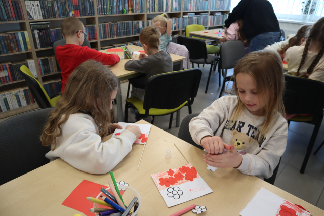 Na zdjęciu znajdują się dziewczynki przy stoliku i przyklejają patyk do swojej flagi. W tle znajdują się inni uczestnicy zajęć oraz półki z książkami.