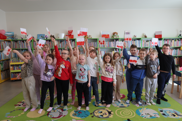 Na zdjęciu znajduje się osiemnaście przedszkolaków stojących do zdjęcia z podniesionymi do góry wykonanymi przez siebie flagami Polski. W tle znajdują się półki z książkami.