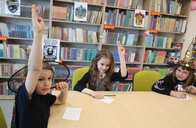 Na zdjęciu znajduje się trójka dzieci przy stoliku i podnoszą rękę do góry. W tle znajdują się półki z książkami.
