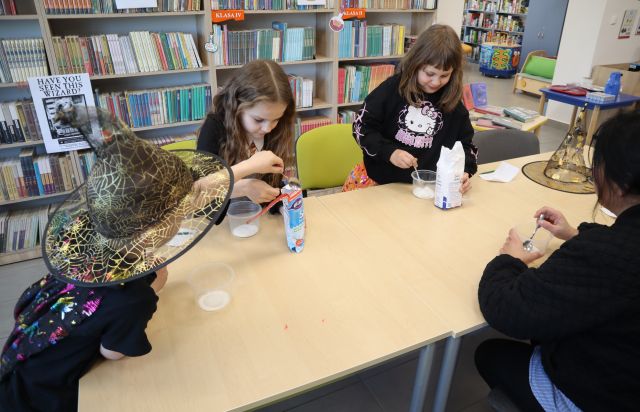 Na zdjęciu znajdują się dwie dziewczynki oraz jeden chłopiec w czarodziejskich strojach przy złączonych stołach i mieszają sodę z cukrem w plastikowych pojemniczkach. Po prawej stronie zdjęcia przy stole siedzi kobieta. W tle znajdują się półki z książkami.
