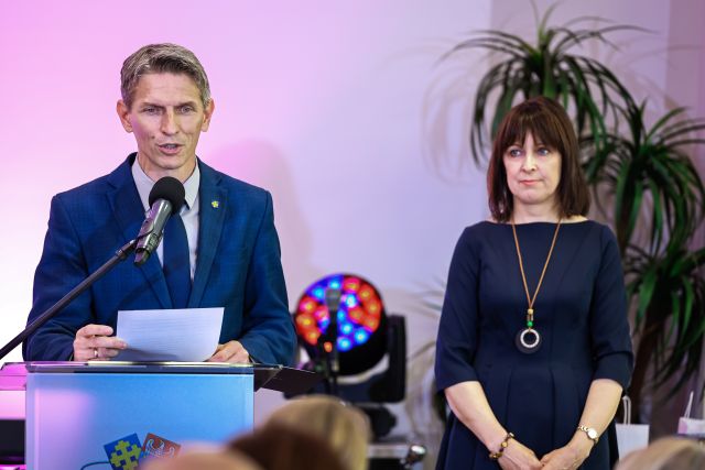 Na zdjęciu znajduje się przemawiający przy mównicy z mikrofonem mężczyzna, a po lewej stronie stoi kobieta, ma złączone dłonie. W tle znajduję się lampa oraz kwiat.