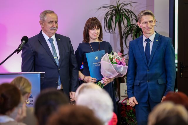 Na zdjęciu znajduje się dwóch mężczyzn, pomiędzy nimi stoi kobieta z teczką oraz bukietem kwiatów, przed nimi siedzą ludzie, a po lewej stronie stoi mównica z mikrofonem. W tle znajduje się palma.