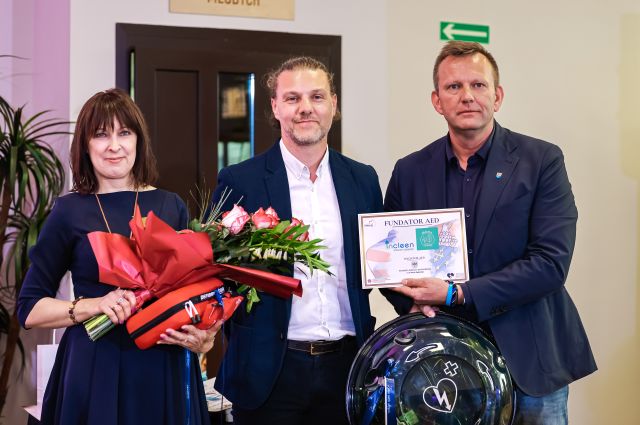 Na zdjęciu znajduje się dwóch mężczyzn oraz jedna kobieta. Kobieta trzyma w rękach kwiaty oraz apteczkę, a mężczyzna po prawej trzyma Fundator AED oraz defibrylator. W tle znajdują się drzwi.