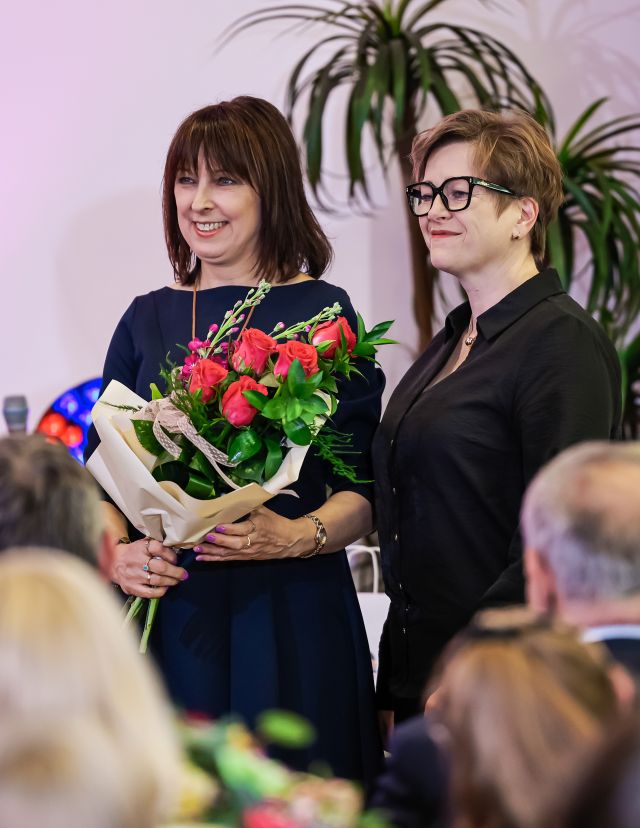 Na zdjęciu znajdują się dwie kobiety stojące wspólnie do zdjęcia, jedna z nich trzyma kwiaty. Za kobietami znajduje się roślina, a przed widownia, uchwycona na zdjęciu od tyłu.