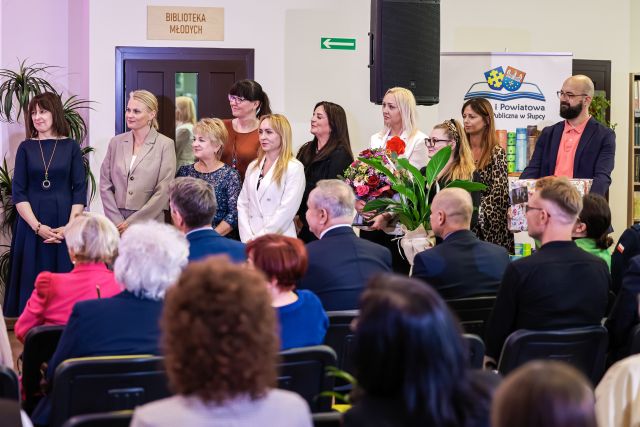 Na zdjęciu znajduje się widownia przed którą stoi dziewięć kobiet oraz jeden mężczyzna. Jedna z kobiet trzyma kwiaty. W tle znajdują się drzwi, głośnik oraz baner z logo.