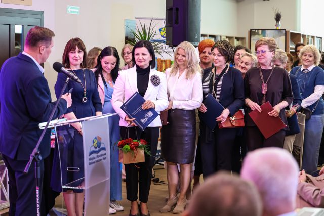 Na zdjęciu znajduje się grupa kobiet oraz jeden mężczyzna stojący przy jednej z kobiet. Po lewej stronie stoi mównica z mikrofonem, a w tle znajduje się głośnik oraz półki z książkami.