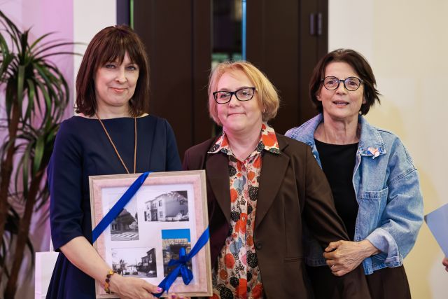 Na zdjęciu znajdują się trzy kobiety stojące do wspólnego zdjęcia. Kobieta stojąca po lewej stronie trzyma w rękach ramkę ze zdjęciami. W tle znajdują się drzwi oraz roślina.