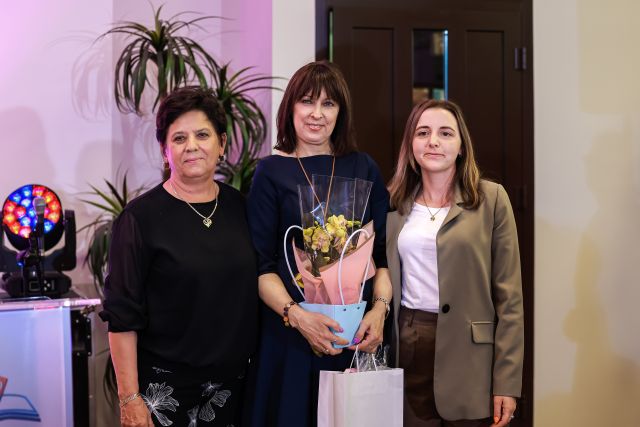 Na zdjęciu znajdują się trzy kobiety stojące razem do zdjęcia, kobieta stojąca w środku trzyma kwiaty. W tle znajdują się drzwi. Roślina oraz lampa.