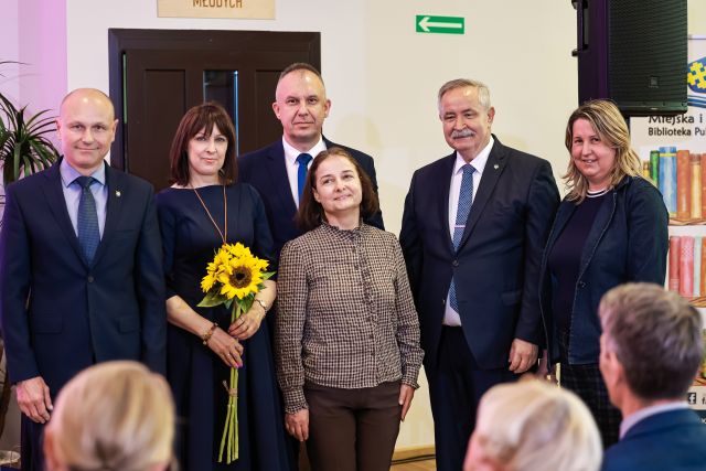 Na zdjęciu znajduje się trójka mężczyzn oraz trzy kobiety, jedna z nich trzyma w ręce słoneczniki. W tle znajdują się drzwi, a po prawej stronie znajduje się cześć banera z logiem i książkami oraz głośnik.
