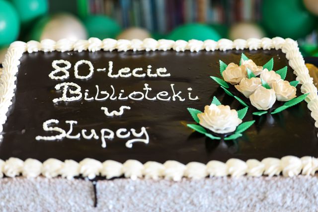 Na zdjęciu znajduje się prostokątny tort z ciemną polewą, znajdują się na nim cztery kwiatki oraz napis 80-lecie Biblioteki w Słupcy.