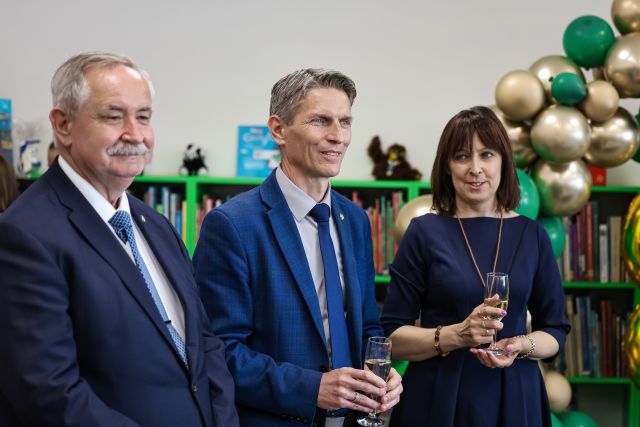 Na zdjęciu znajduje się dwóch mężczyzn oraz kobieta., w ręce trzymają szampana,, a w tle znajdują się półki z książkami oraz balony.