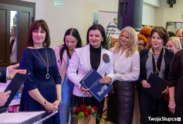 Na zdjęciu znajduje się grupa kobiet, dwie z nich trzymają teczkę. Po lewej stronie znajduje się mównica, przy której stoi mężczyzna, na zdjęciu znajdują się tylko jego ręcę i trzymana przez niego teczka. W tle znajduje się głośnik, baner z logiem biblioteki.