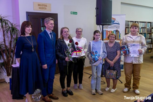 Na zdjęciu znajdują się cztery kobiety, jeden mężczyzna, chłopiec oraz dziewczynka. W rękach trzymają książki. W tle znajdują się drzwi, głośnik oraz półki z książkami.