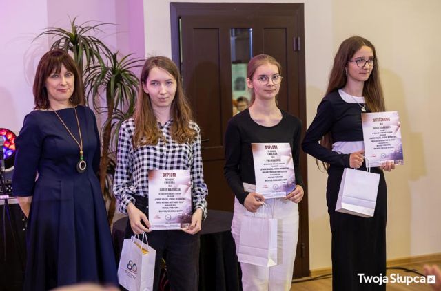 Na zdjęciu znajduje się kobieta oraz trzy nastolatki z dyplomami i upominkami. W tle znajdują się drzwi oraz kwiat.