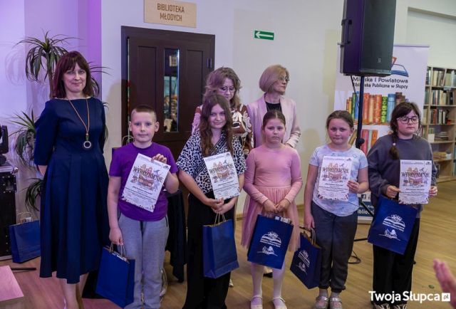 Na zdjęciu znajdują się trzy kobiety oraz piątka dzieci z dyplomami i upominkami. W tle znajdują się drzwi, głośnik oraz baner z logiem biblioteki i książkami.