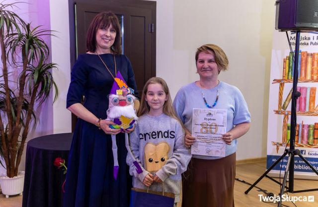 Na zdjęciu znajdują się dwie kobiety i dziewczynka pomiędzy nimi. Kobieta po lewo trzyma maskotkę, krasnala, kobieta po prawo dyplom, a dziewczynka upominek. W tle znajdują się drzwi, sztuczne drzewko oraz baner z logiem biblioteki i książkami po prawej stronie.