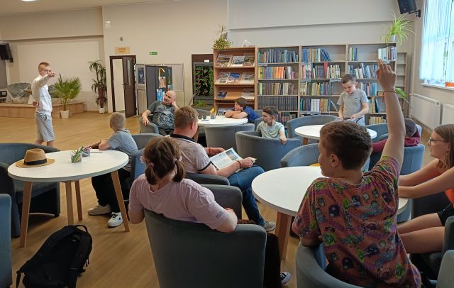 Na zdjęciu znajduje się młodzież przy okrągłych stolikach, po lewej stronie na holu biblioteki stoi jedna osoba. W tle znajdują się półki z książkami.