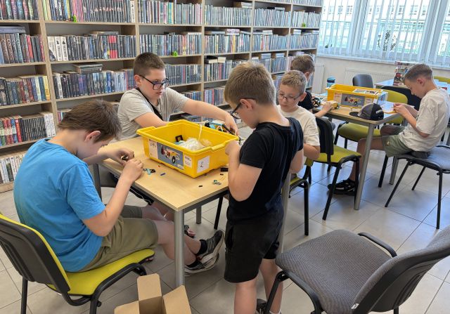 Na zdjęciu znajduje się szóstka chłopaków przy dwóch stołach i układają znajdujące się na stolikach klocki LEGO. W tle znajdują się półki z książkami oraz okna po prawej stronie.