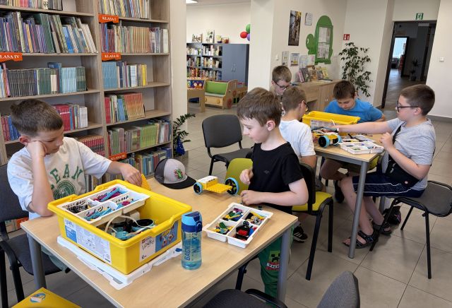 Na zdjęciu znajduje się dwóch chłopców przy stoliku i układają znajdujące się na nim klocki LEGO, po prawej stronie przy osobnym stoliku siedzi czwórka chłopców i również układają LEGO. W tle znajdują się półki z książkami oraz wejście do innej części z książkami.