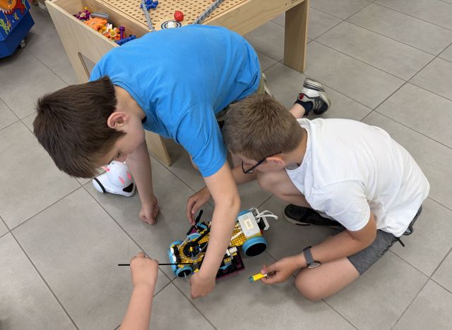 Na zdjęciu znajduje się dwójka chłopców, którzy kucają i poprawiają coś w pojeździe z LEGO, który stoi na podłodze. Obok jednego chłopca znajduje się robot Photon oraz stolik edukacyjny.